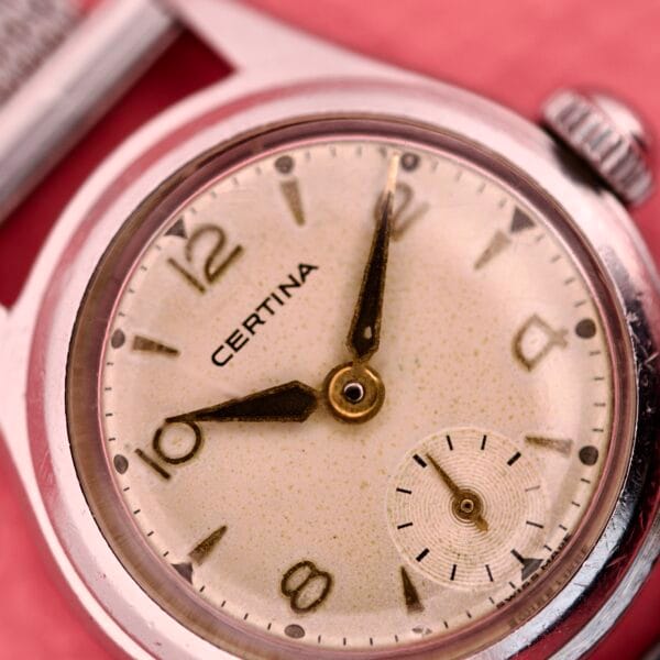 Close-up of a Ladies Certina Ref. 41101 wristwatch in stainless steel, featuring gold hands, Cal. 19-10 movement, and a small seconds dial, set against a pink background.