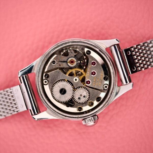Close-up of the Ladies Certina Ref. 41101 in stainless steel with exposed Cal. 19-10 mechanical gears, displayed on a pink textured background.