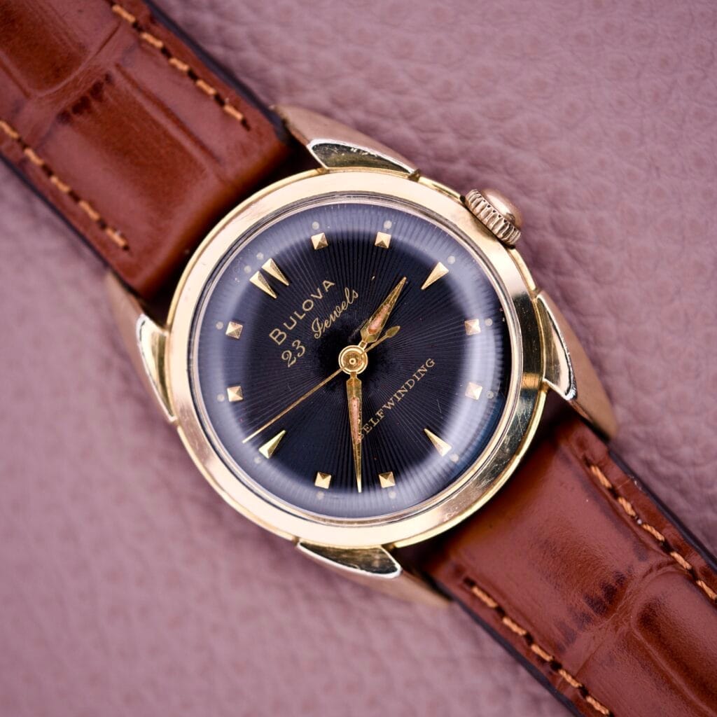 1957 Bulova 23 Jewels Self Winding Cal. 10BPAC gold wristwatch with brown leather strap shown on a textured pink surface.