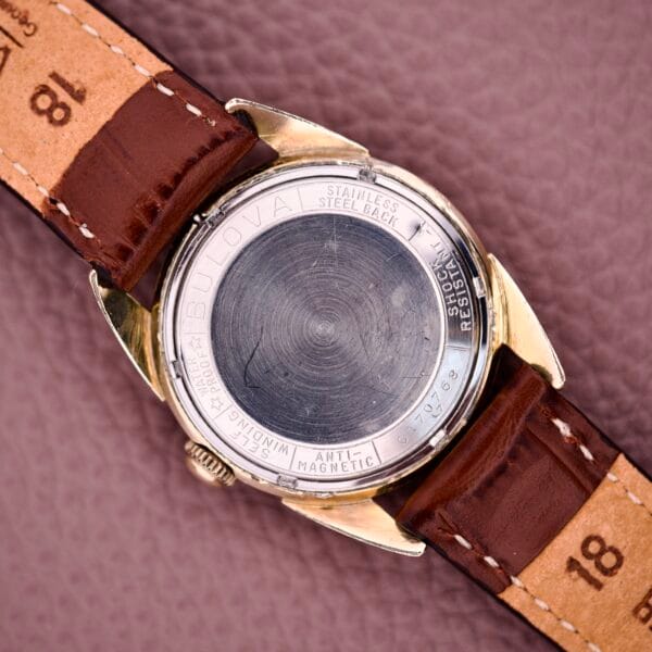 Close-up of the back of a 1957 Bulova 23 Jewels Self Winding Cal. 10BPAC watch with brown leather straps on a textured surface, highlighting its self-winding movement.