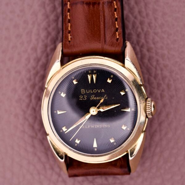 A 1957 Bulova 23 Jewels Self Winding Cal. 10BPAC wristwatch with a brown leather strap displayed on a textured pink background.