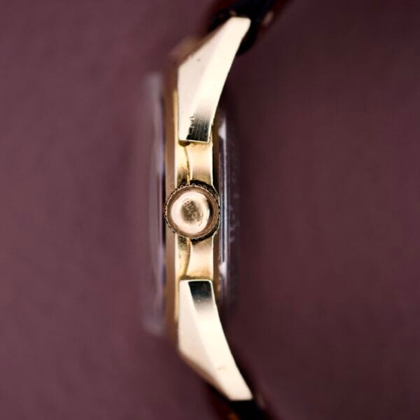 Close-up side view of the 1957 Bulova 23 Jewels Self Winding Cal. 10BPAC gold case with crown and brown leather strap on a mauve background, highlighting its self-winding movement and 23-jewel marking.