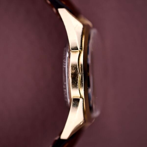 Close-up side view of the 1957 Bulova 23 Jewels Self Winding Cal. 10BPAC wristwatch with a gold case and brown leather strap, set against a pink background.