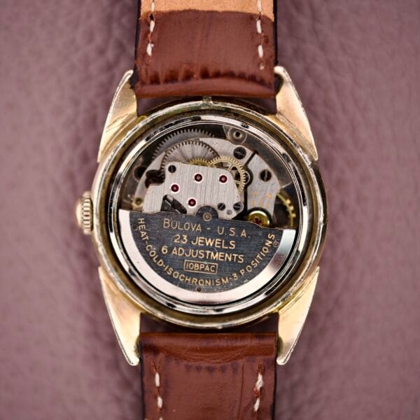 Close-up of a 1957 Bulova 23 Jewels Self Winding Cal. 10BPAC watch movement with visible gears, engraved 23 Jewels and Self Winding, shown on a brown leather strap.