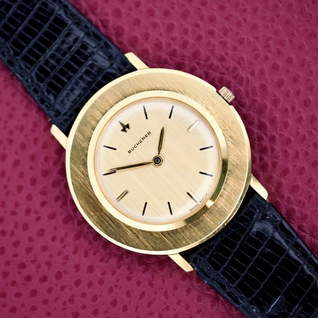 Bucherer Disco Volante Thin Manual Wind Caliber 280 wristwatch with a black leather strap displayed on a textured pink surface.