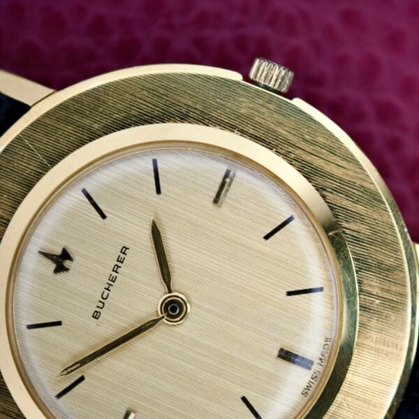 Close-up of a gold Bucherer Disco Volante Thin Manual Wind Caliber 280 wristwatch with a textured face and black hour markers on a pink background, showcasing its elegant manual wind mechanism.