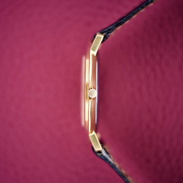 Side view of the Bucherer Disco Volante Thin Manual Wind Caliber 280 gold wristwatch with a black strap, displayed against a textured pink background.