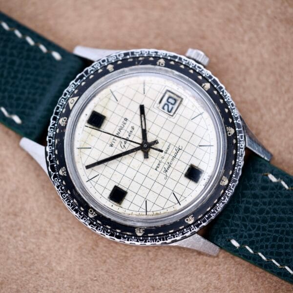 The Wittnauer Pert-O-Graph Ref. 7005 features a grid-patterned white dial, rare Pert-O-Graph Scale Bezel, and green leather strap, displayed on a tan background.