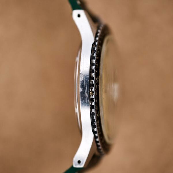 Side view of a Wittnauer Pert-O-Graph Ref. 7005 wristwatch featuring a scratched metal case and its rare Pert-O-Graph scale bezel, worn and chipped, set against a brown background.