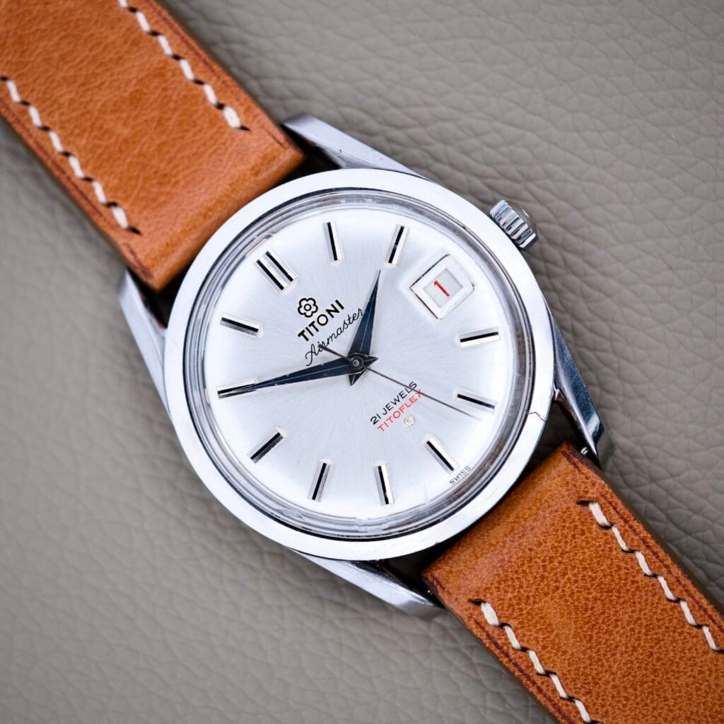 A Titoni Air Master 305-345 wristwatch featuring a silver case, brown leather strap, date display, black hour markers, and a stainless steel 21 Jewels Titoflex movement.