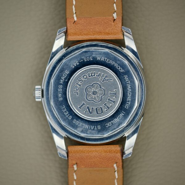 Close-up of the Titoni Air Master 305-345 wristwatch with a tan leather strap, stainless steel case, and 21 jewels Titoflex movement, displayed on a gray surface.