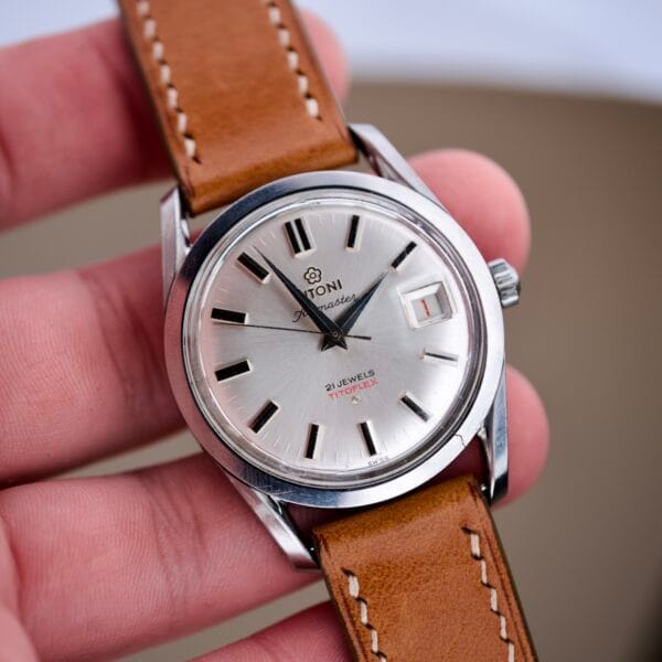 A hand holds a Titoni Air Master 305-345 watch with a stainless steel case, brown leather strap, silver dial, and date display; features Titoflex and 21 jewels.