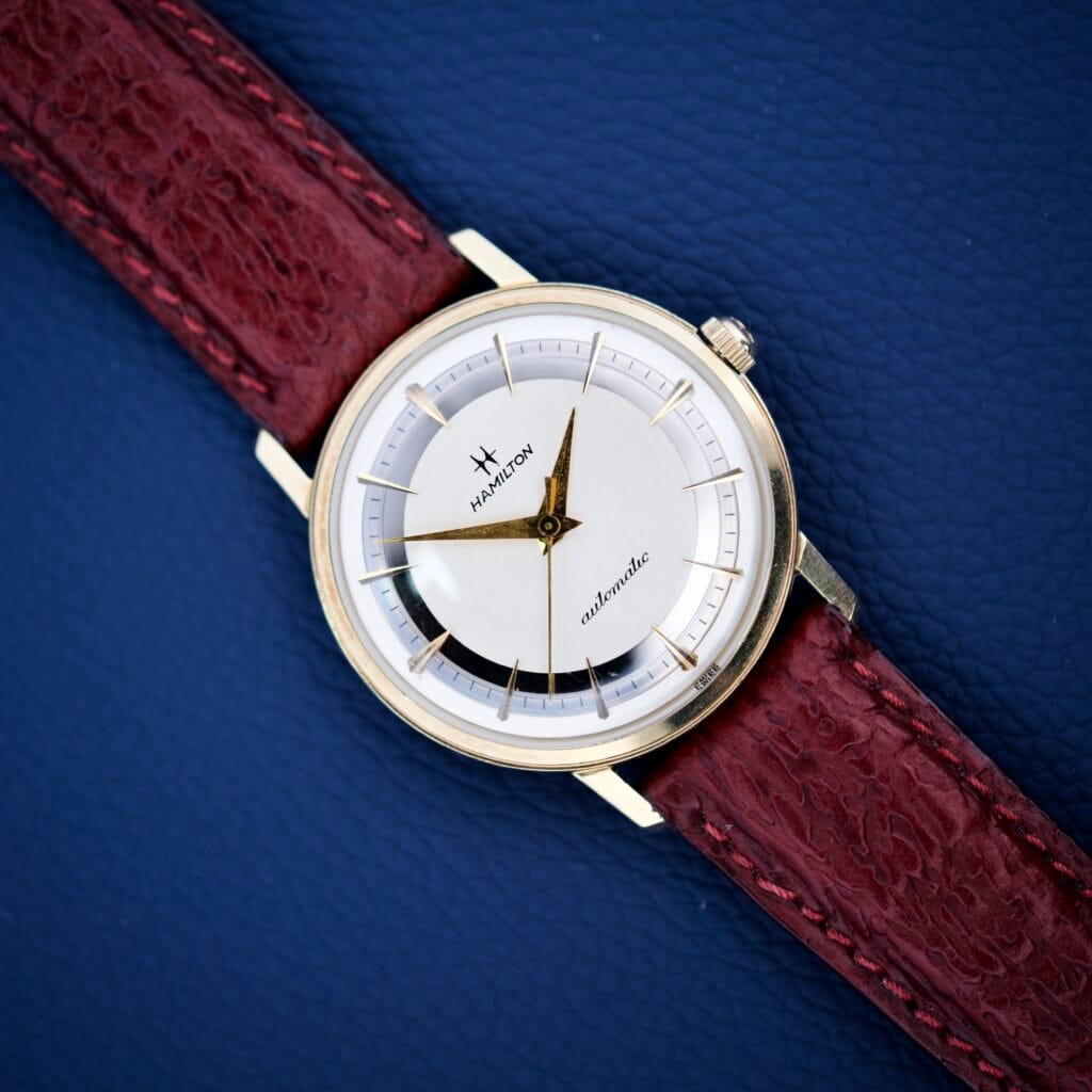 A 1964 Hamilton Accumatic A-602 Silver Chapter Ring Dial Cal. 669 wristwatch with a white face and red leather strap rests stylishly on a textured blue surface.