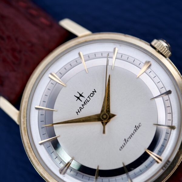 Close-up of a 1964 Hamilton Accumatic A-602 with Silver Chapter Ring Dial, Cal. 669 movement, brown leather strap, and gold accents displayed on a blue background.