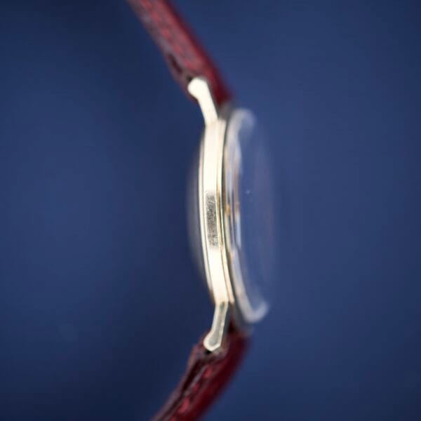 Side view of a 1964 Hamilton Accumatic A-602 wristwatch with a round case, Silver Chapter Ring Dial Cal. 669, and red leather strap, set against a blue background.