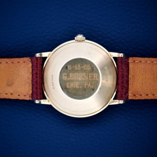 The 1964 Hamilton Accumatic A-602 with silver chapter ring dial and Cal. 669, features a gold engraved back (6-13-65 G. Bosner Erie, PA.), brown leather strap, and is displayed on a blue surface for an elegant presentation.