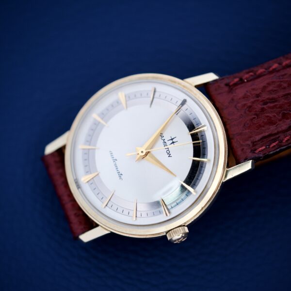 The 1964 Hamilton Accumatic A-602 with a Silver Chapter Ring Dial and brown leather strap rests on a blue textured background.
