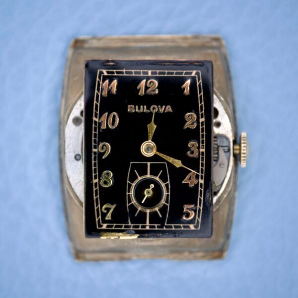 Close-up of a Bulova Art Deco Black Dial With Arabic Numerals Cal. 10 AK wristwatch, featuring gold numerals, displayed against a light blue background.