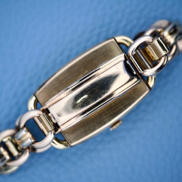 Close-up of a gold metal bracelet clasp against a textured light blue background, inspired by the Bulova Art Deco Black Dial With Arabic Numerals Cal. 10 AK.