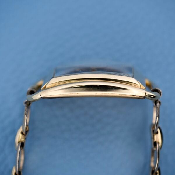 The Bulova Art Deco Black Dial With Arabic Numerals Cal. 10 AK features a gold curved rectangular case, black dial with Arabic numerals, and a chain band, displayed from the side on a blue background.