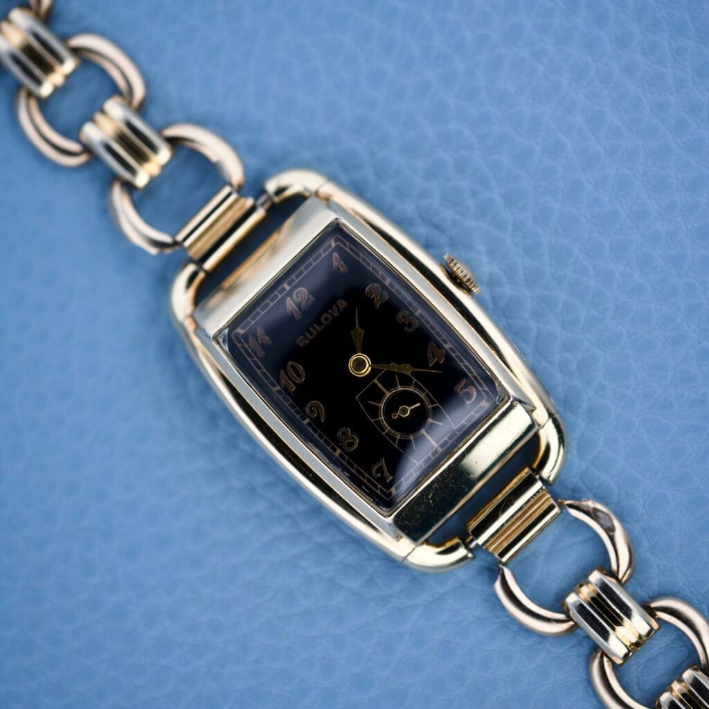 Bulova Art Deco Black Dial With Arabic Numerals Cal. 10 AK wristwatch with a rectangular face, shown on a blue textured background.