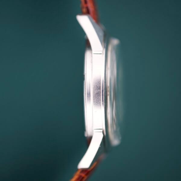 Side view of the Gruen Precision White Dial From France Cal. 515SS silver wristwatch with brown leather strap and white dial, set against a dark green background.