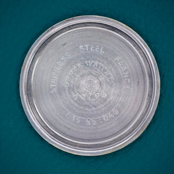 Round stainless steel case back of the Gruen Precision White Dial From France Cal. 515SS, featuring engraved text, displayed on a teal textured surface.
