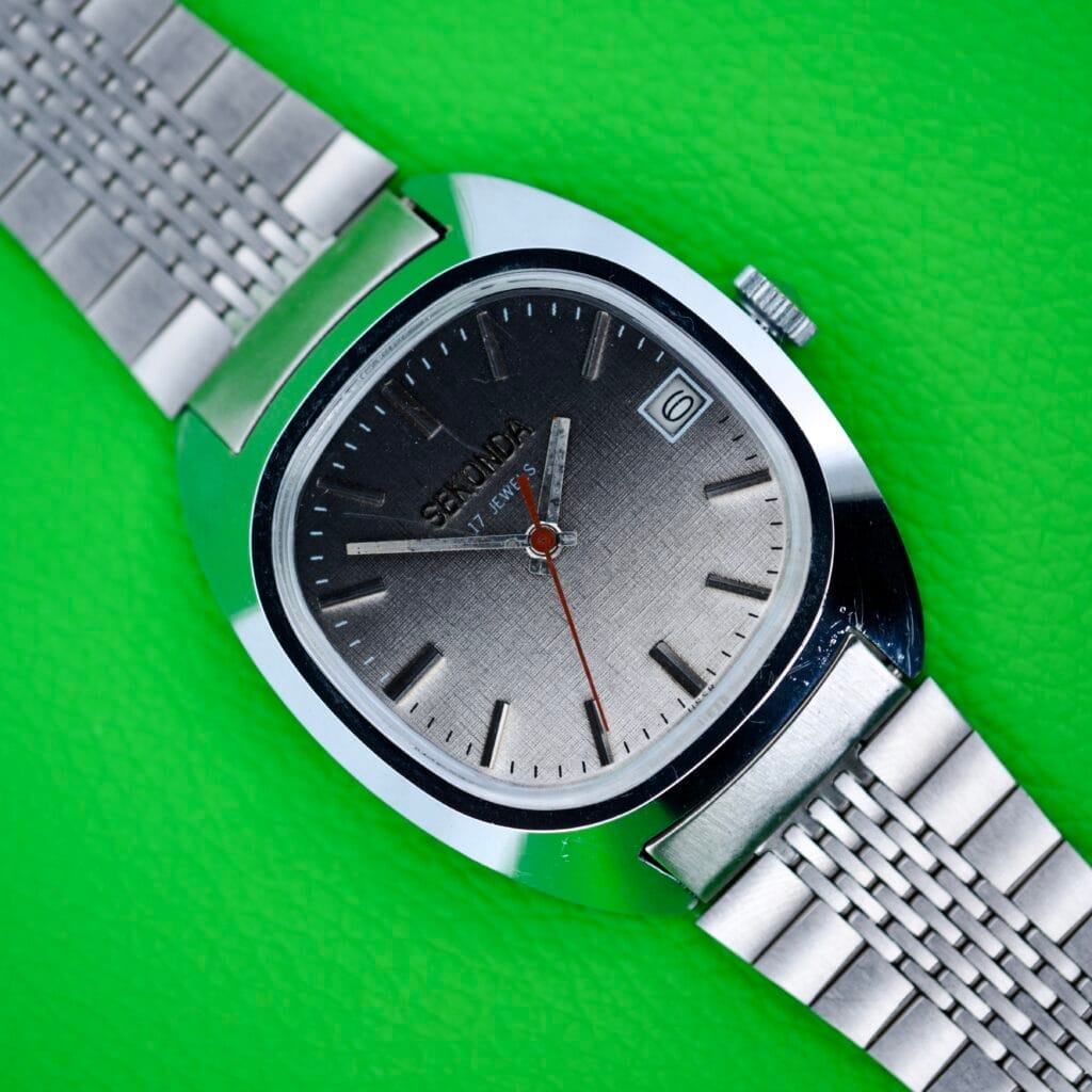 Seconda Ombre Grey Dial In Stainless Steel Cal. 2614 2H wristwatch with a metal band and ombre grey dial, set to 10:10, displayed on a bright green textured background.