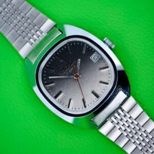 Seconda Ombre Grey Dial In Stainless Steel Cal. 2614 2H wristwatch with a metal band and ombre grey dial, set to 10:10, displayed on a bright green textured background.