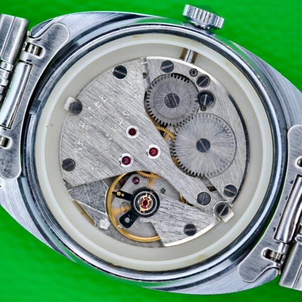 Close-up of the Seconda Ombre Grey Dial in Stainless Steel, showcasing its Cal. 2614 2H mechanical movement with visible gears and springs, set against a bright green background.