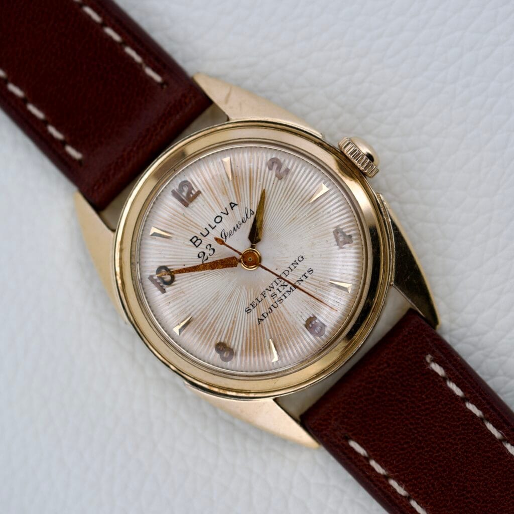 The 1965 Bulova 23 Jewels Six Adjustments Automatic features a gold case, brown leather band, white textured dial, and displays the time at 8:22.