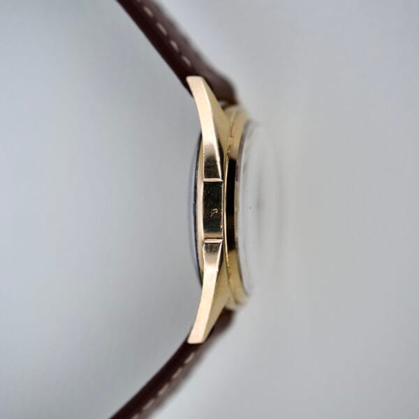 Side view of a 1965 Bulova 23 Jewels Six Adjustments Automatic gold wristwatch case with a brown leather strap, featuring a small hallmark on the edge. This automatic watch offers precision with its 23 jeweled movement.
