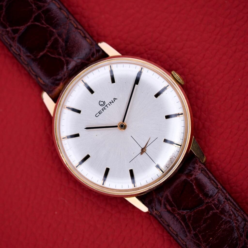 Certina Ref. 7400-207 wristwatch with a rose gold tone case, silver dial, and brown leather strap, Cal. 28-10, displayed on a textured red surface.