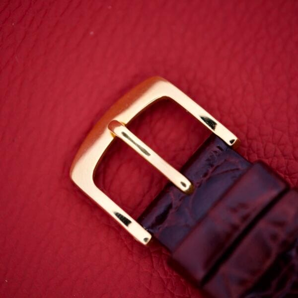 Close-up of a Certina Ref. 7400-207 Rose Gold Tone Case Cal. 28-10 buckle, attached to a dark brown leather strap on a red textured surface.
