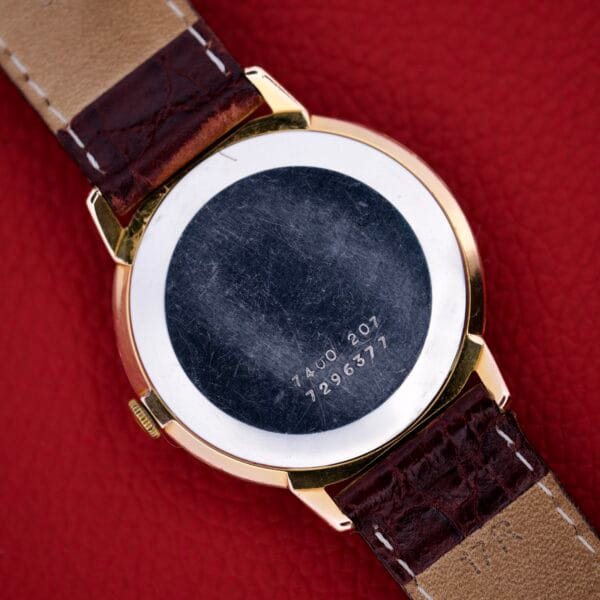 The back of a Certina Ref. 7400-207 Rose Gold Tone Case Cal. 28-10 with a brown leather strap and engraved serial numbers rests on a red textured surface.