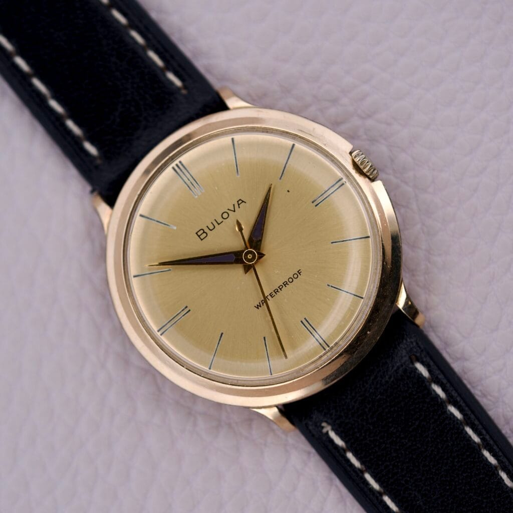 The Bulova Gold Sunburst Dial & Case Cal. 11AFC wristwatch showcases a stunning gold sunburst dial with a black leather strap, elegantly displayed against a light textured background.