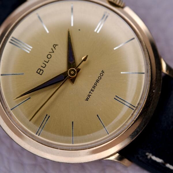 Close-up of a Bulova Gold Sunburst Dial & Case Cal. 11AFC wristwatch, featuring a gold sunburst dial, black band, and WATERPROOF inscribed on the face.