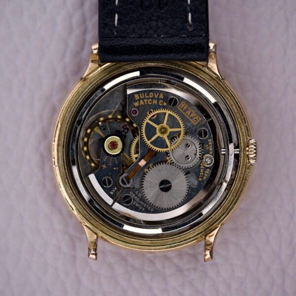 Close-up of the Bulova Gold Sunburst Dial & Case Cal. 11AFC watch, showcasing its exposed mechanical movement and visible gears, resting on a textured surface.