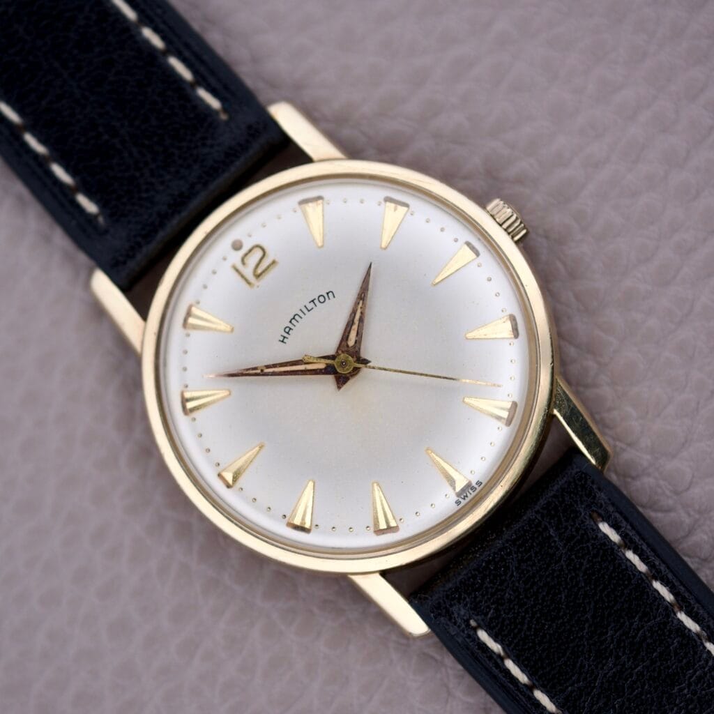 A Hamilton Sea-Scape Sunburst Dial Manual Wind Cal. 671 wristwatch with a gold case and black leather strap rests on a textured surface.