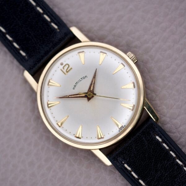A Hamilton Sea-Scape Sunburst Dial Manual Wind Cal. 671 wristwatch with a gold case and black leather strap rests on a textured surface.