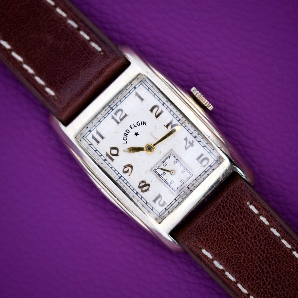 A 1936 Lord Elgin Star Dial Tank Style Case Manual Wind Grade 531 wristwatch with a brown leather strap rests on a purple textured surface.