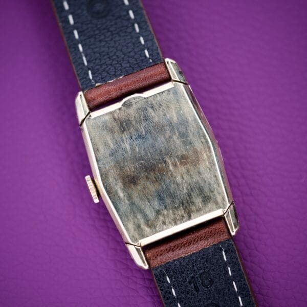 A 1936 Lord Elgin Star Dial Tank Style Case Manual Wind Grade 531 wristwatch, with a worn metal case and black leather strap, rests on a purple textured surface, showcasing classic Tank Style elegance.