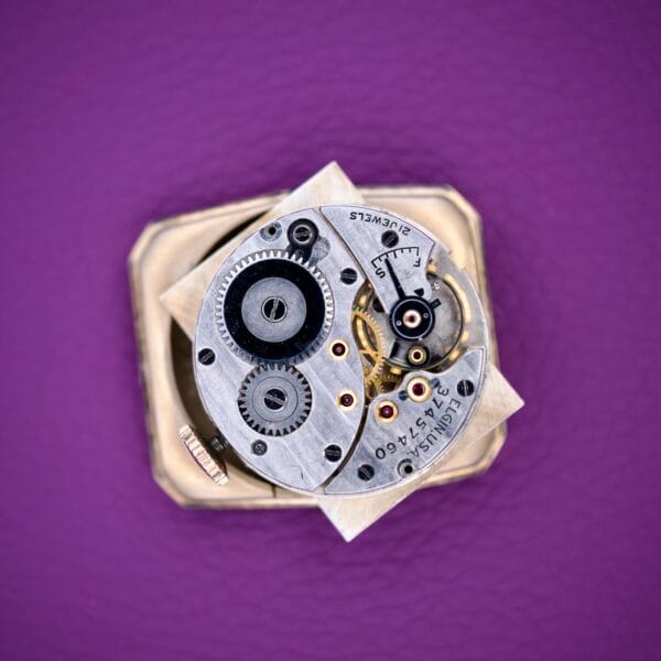 Close-up of a 1936 Lord Elgin Star Dial Tank Style Case Manual Wind Grade 531 watch mechanism with gears, set against a purple textured background.