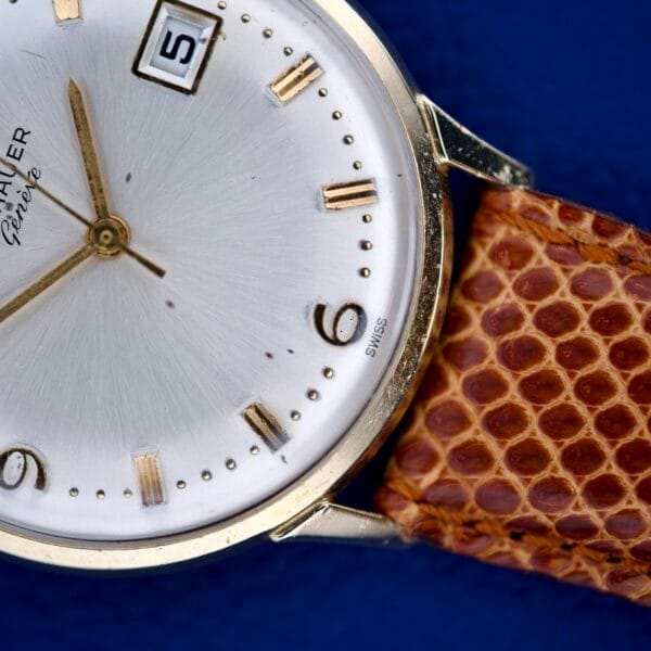 A close-up of the Wittnauer Geneve Manual Wind Date & Sunburst Dial watch, featuring a gold case, sunburst dial, brown textured strap, and date display, set against a blue background.