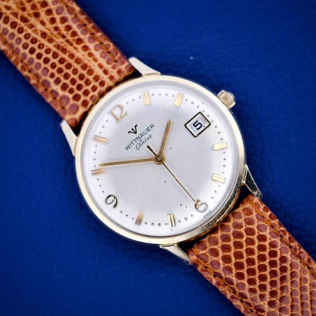 A Wittnauer Geneve Manual Wind watch with date and sunburst dial on a brown leather strap is displayed against a blue background.