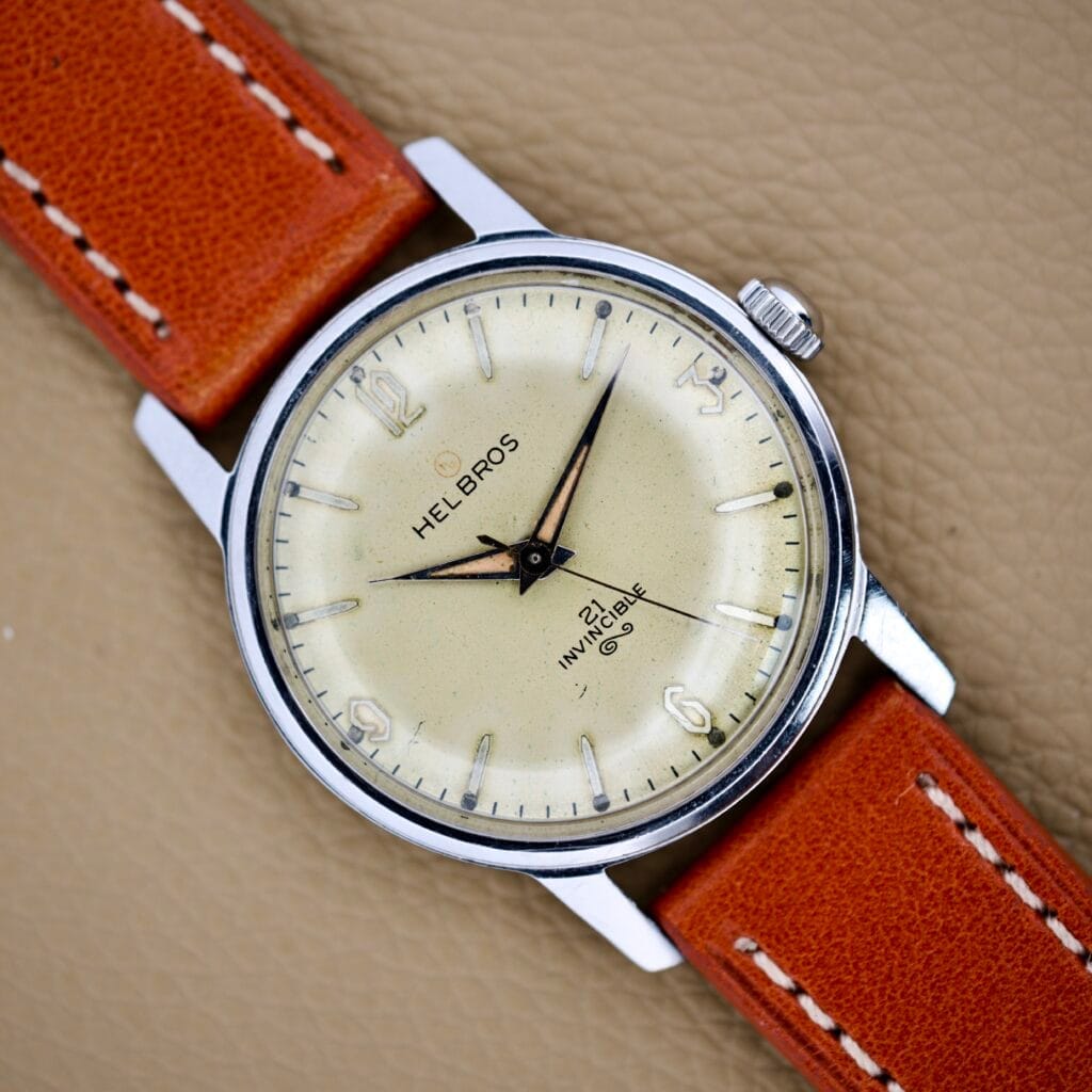 Helbros Invincible 21 Jewels Manual Wind wristwatch with a cream dial, orange leather strap, and stainless steel case, displayed on a tan textured surface.