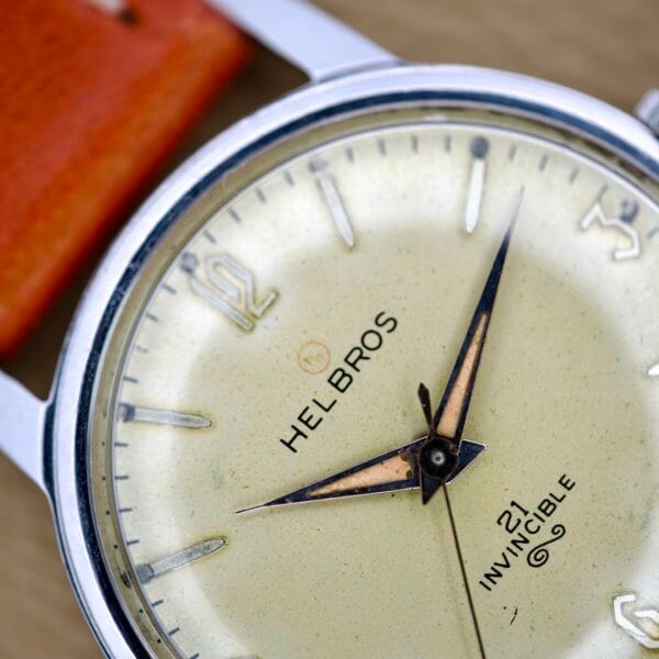 Close-up of a Helbros Invincible 21 Jewels wristwatch with a tan leather strap, gold hands at 8:22, and a stainless steel case. This classic timepiece features precise manual wind movement.