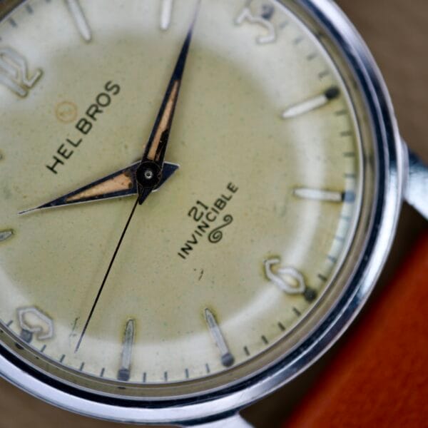 Close-up of a Helbros Invincible 21 Jewels Manual Wind wristwatch with a stainless steel case, cream dial, and brown leather strap.