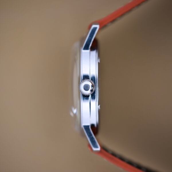 Side view of a Helbros Invincible 21 Jewels Manual Wind watch with stainless steel case, silver crown, and orange strap, set against a plain brown background.