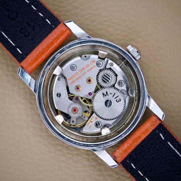 Close-up of a Helbros Invincible 21 Jewels Manual Wind watch with an exposed movement, stainless steel case, and a red and black strap displayed on a tan textured surface.